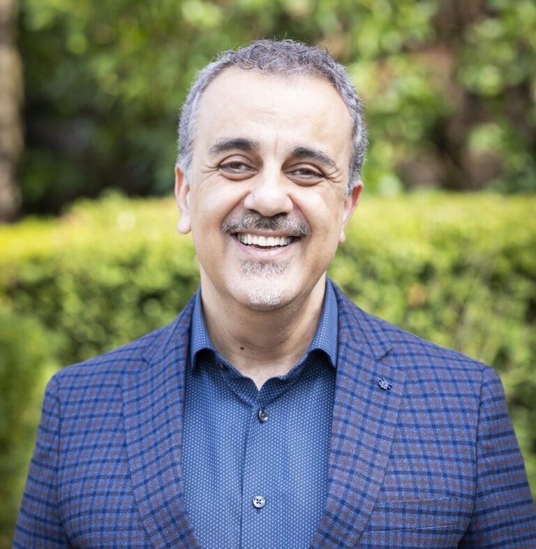 Smiling middle-aged man in a blue checked blazer standing outdoors against greenery