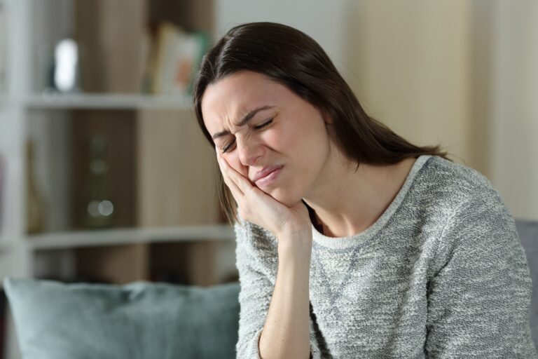 Woman wincing and holding her cheek, suggesting tooth or jaw pain indoors