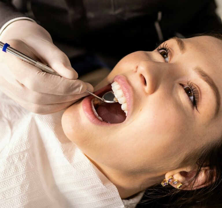 Dentist examines a woman’s teeth with a mirror and dental tool while she reclines in a chair