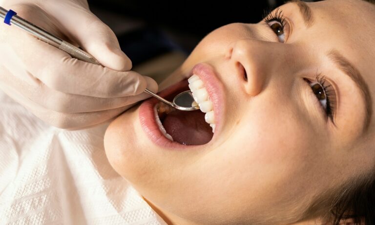 Dentist examines a woman’s teeth with a mirror and dental tool while she reclines in a chair