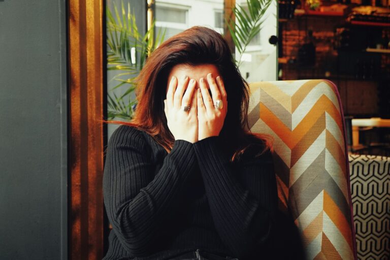 Person sitting in a café chair with hands covering their face, suggesting stress or embarrassment