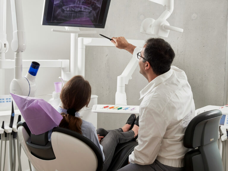 Dentist points to a panoramic dental X-ray on a monitor while a patient sits in the exam chair