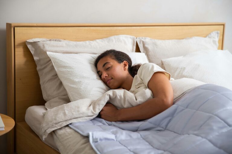 Person sleeping peacefully in bed under a light blue weighted blanket with white pillows behind them