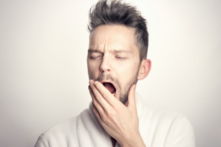 Tired man in a white robe yawning with his hand over his mouth against a plain background