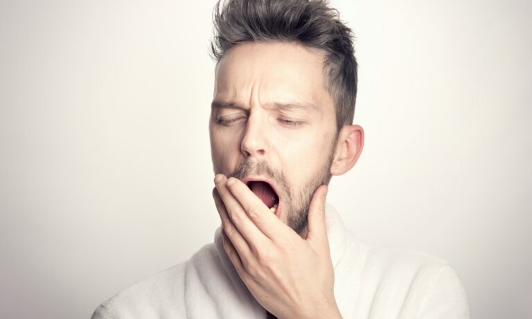 Tired man in a white robe yawning with his hand over his mouth against a plain background
