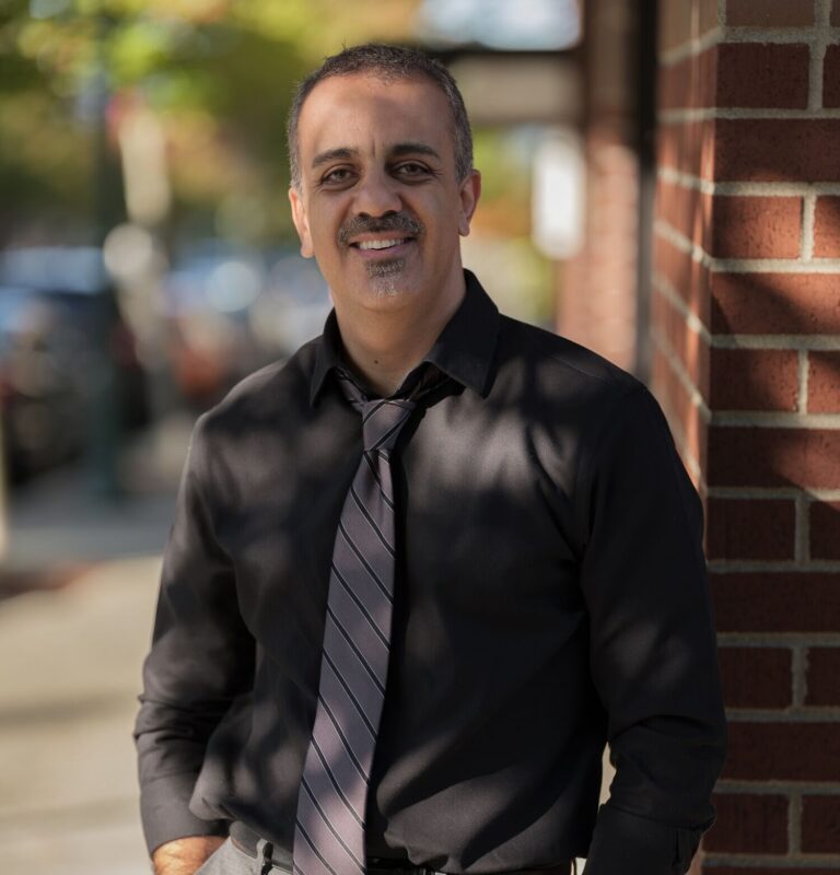Smiling man in a black shirt and striped tie standing beside a brick wall outdoors