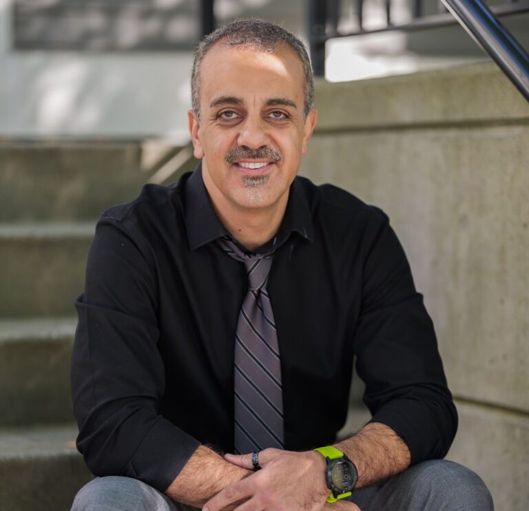 Smiling man in a black shirt and striped tie seated on stairs outdoors