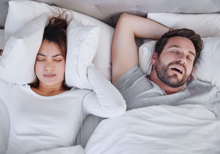 Woman covering her ears with pillows while two men sleep beside her in bed