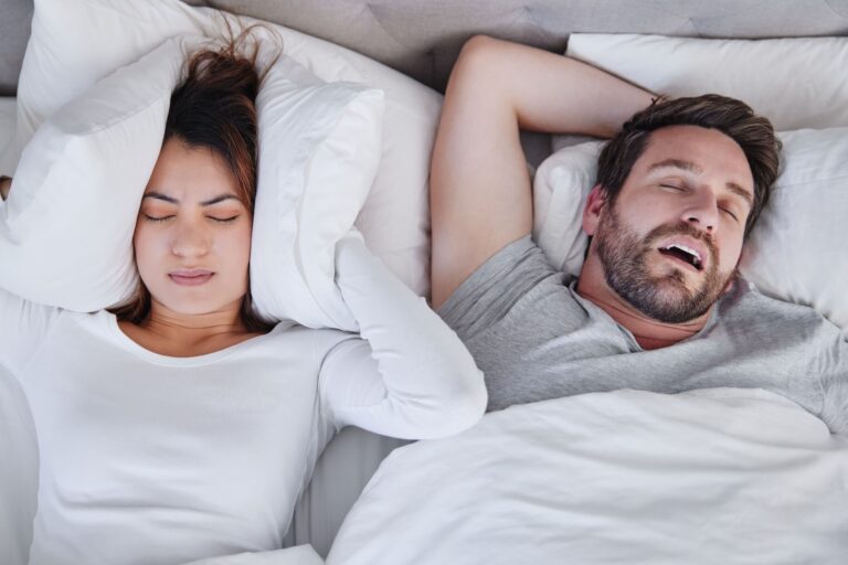 Woman covering her ears with pillows while two men sleep beside her in bed