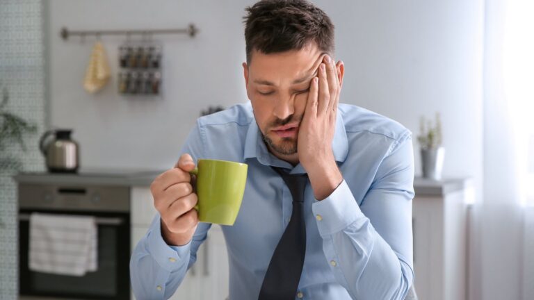 Man in a dress shirt and tie resting his head on his hand, eyes closed, holding a mug of coffee at a kitchen table in the morning.