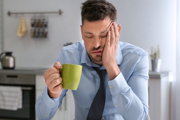 Man in a dress shirt and tie resting his head on his hand, eyes closed, holding a mug of coffee at a kitchen table in the morning.