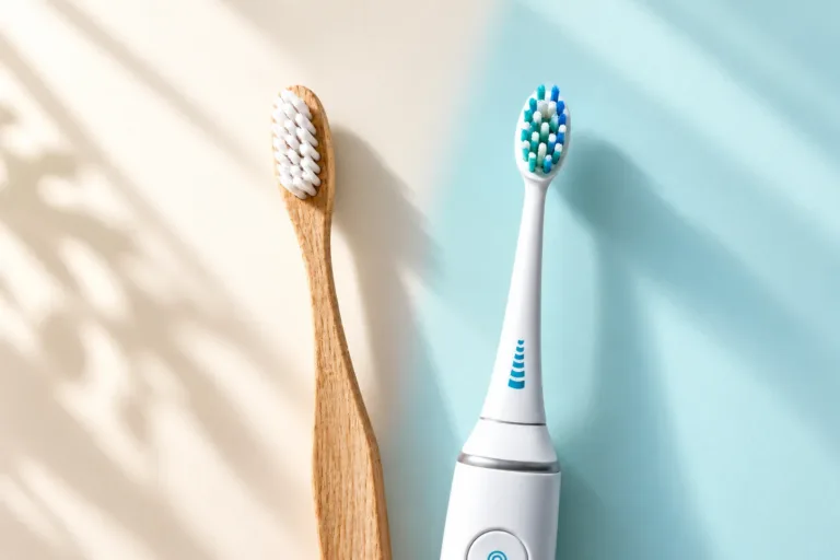 Bamboo manual toothbrush beside a white electric toothbrush on a cream and light-blue background with soft leaf shadows.