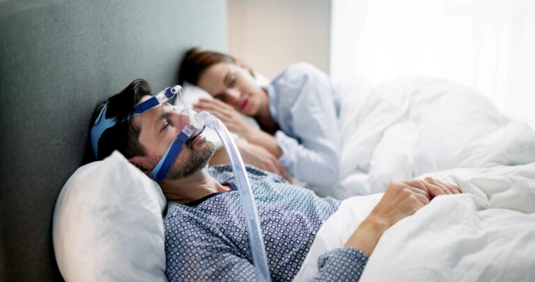 A man sleeps wearing a nasal CPAP mask and tubing while his partner sleeps beside him.