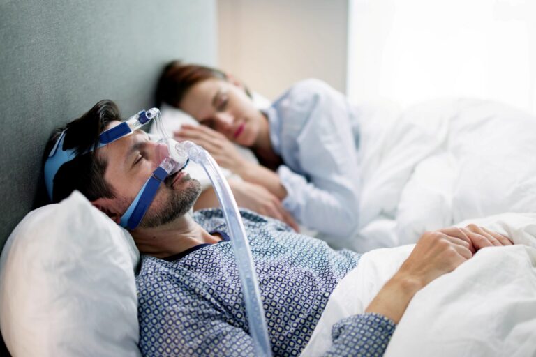 A man sleeps wearing a nasal CPAP mask and tubing while his partner sleeps beside him.